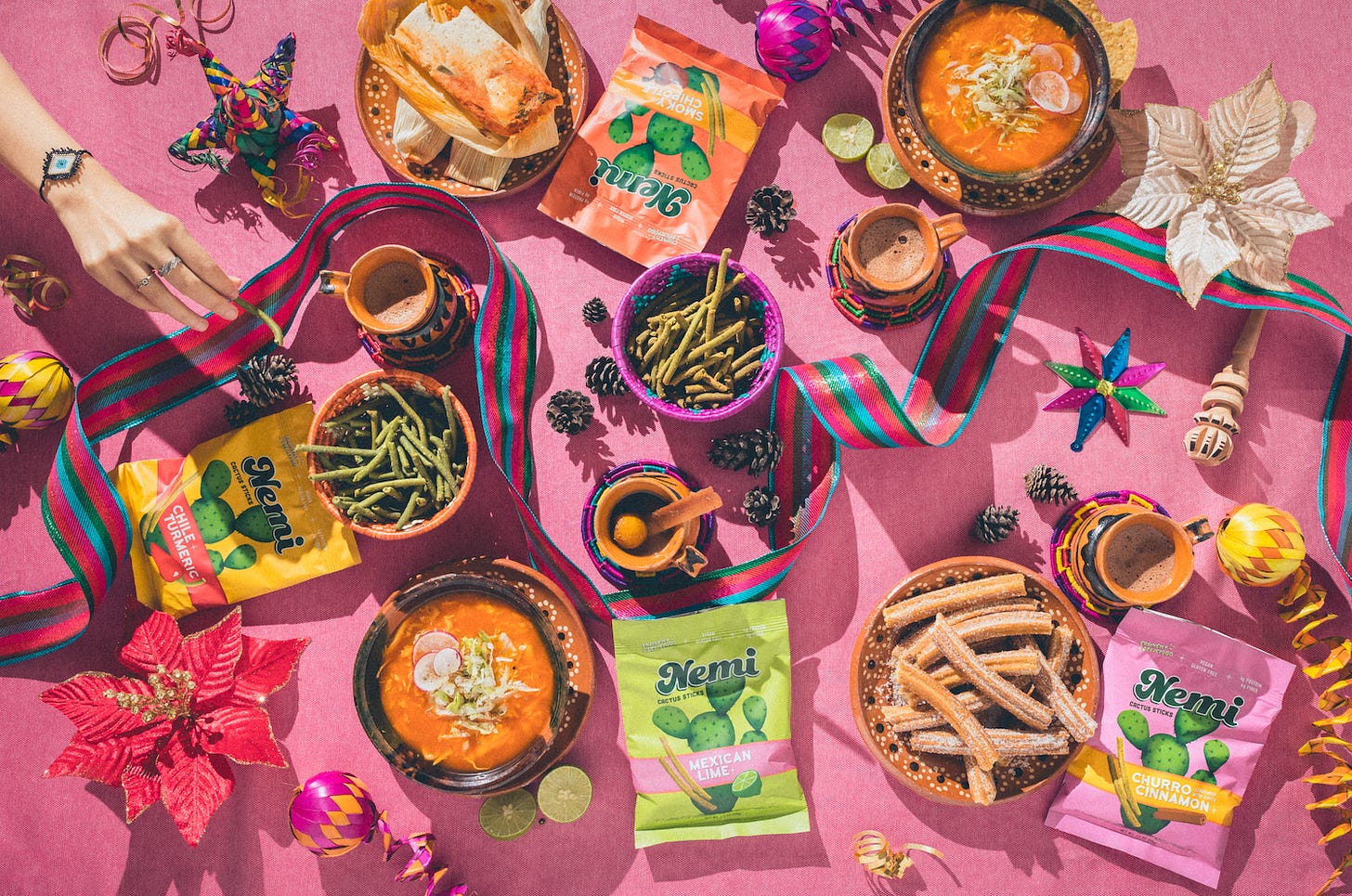 Colorful table with bags of Nemi Snacks nopales snack bags in chile-turmeric, Mexican-lime, smoky-chipotle, and, churro-cinnamon flavors surrounded by Mexican food and dips on a pink tablecloth surrounded by brightly colored traditions Mexican decorations.