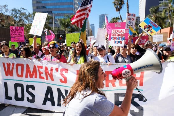 A crowd marching behind a banner that says “Women’s March Los Angeles.” People in the crowd hold up signs with slogans like “Trump is a traitor” and “Bans off our bodies.”