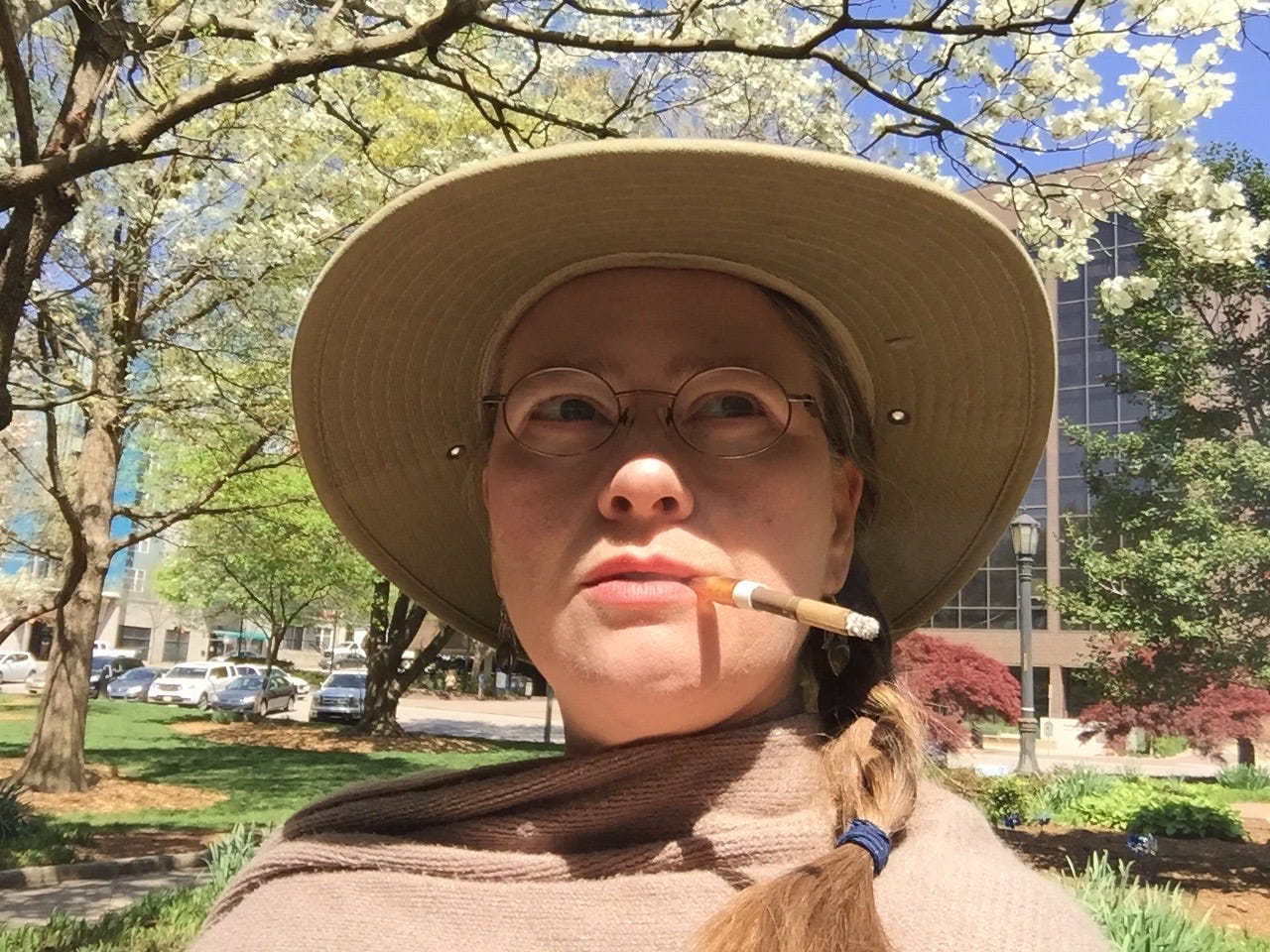 Phot of Jennifer Cerutti in a broad rimmed hat, wearing braids, round eyeglasses, with a lit cigarette hanging from her lips. Behind her dogwood trees are blooming with bright white flowers.