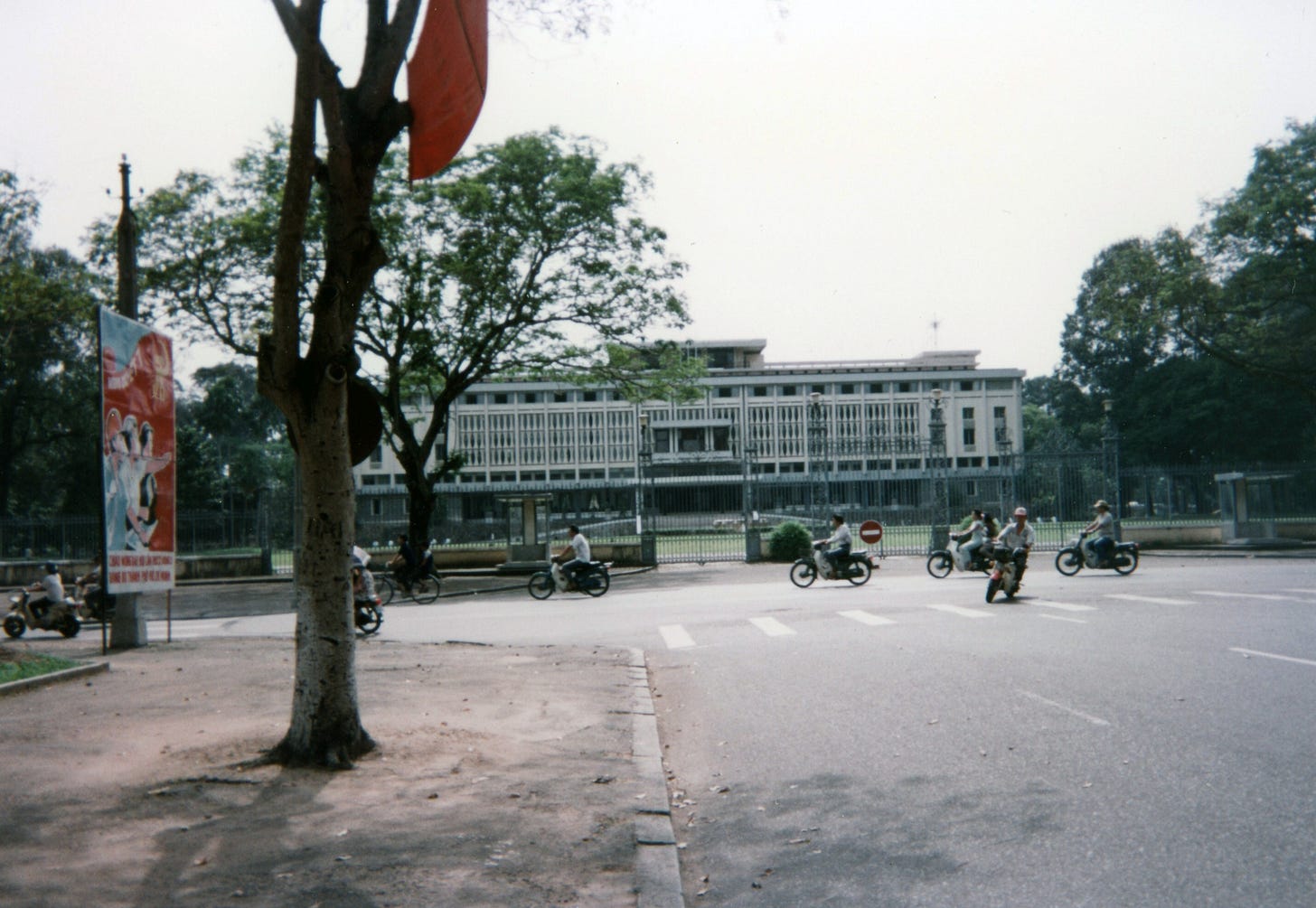A low white building behind a huge fence is set back from the street. There are scooters passing on the road in front of it. The photo is grey and faded.