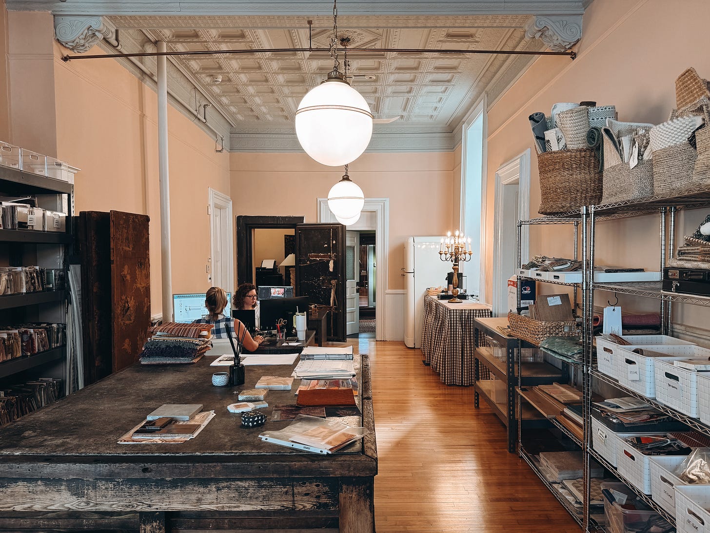 Interior of a design studio with high ceilings and ornate molding, featuring shelves of baskets and samples, two designers working at computers, and a large rustic wood table covered with swatches, drawings, and tools.