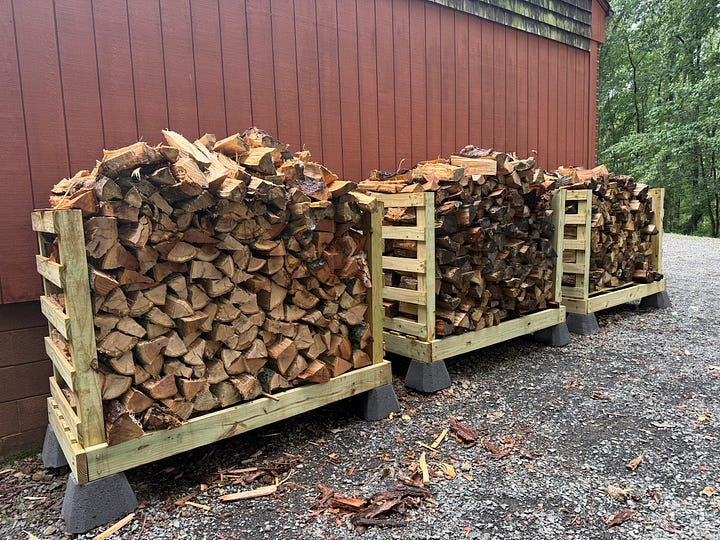 Two side-by-side photos showing a homemade firewood rack before and after being filled. The left image shows a newly built wooden storage rack mounted on concrete deck blocks beside a red barn wall. The right image shows the completed rack packed with neatly stacked split firewood, with additional stacks extending beyond the rack. The photos illustrate the transformation from an empty firewood storage structure to a fully stocked woodshed.
