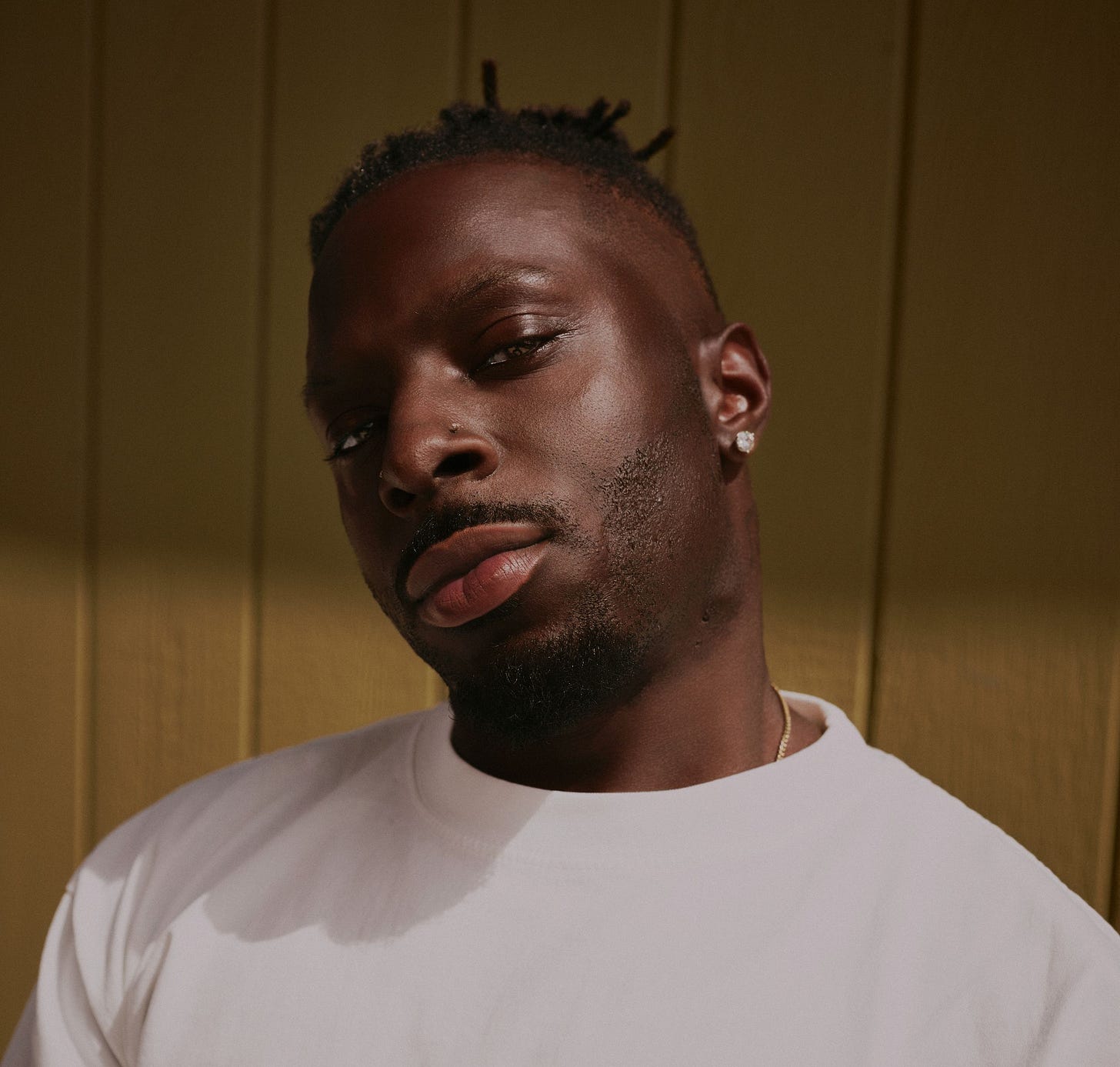 Isaiah Rashad in a white t shirt looking at the camera in front of a green wall.