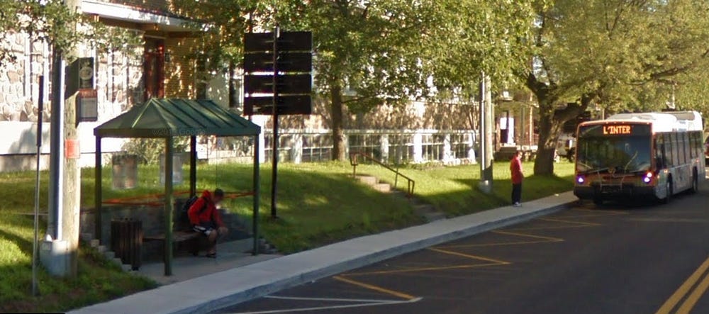 An urban roadway with buildings in the background, sidewalks, and a transit shelter with a bus in the background.