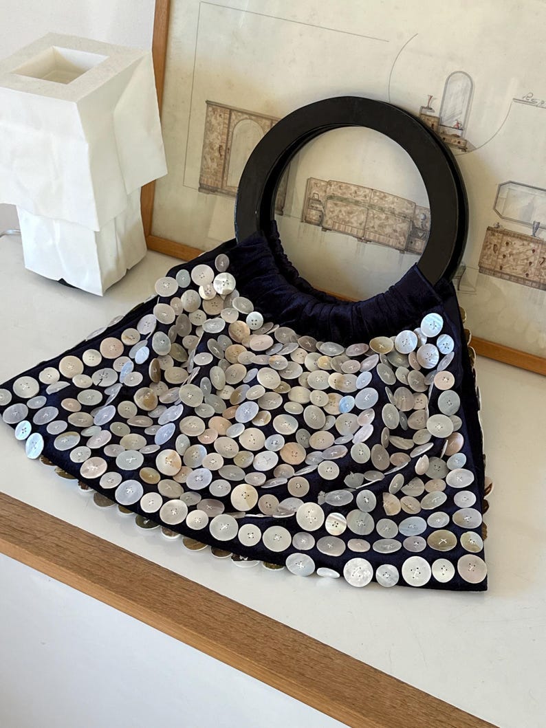 May include: A navy blue handbag with a large black circular handle. The bag is covered in numerous white buttons. The bag is resting on a white surface, with a white paper object and framed artwork in the background.