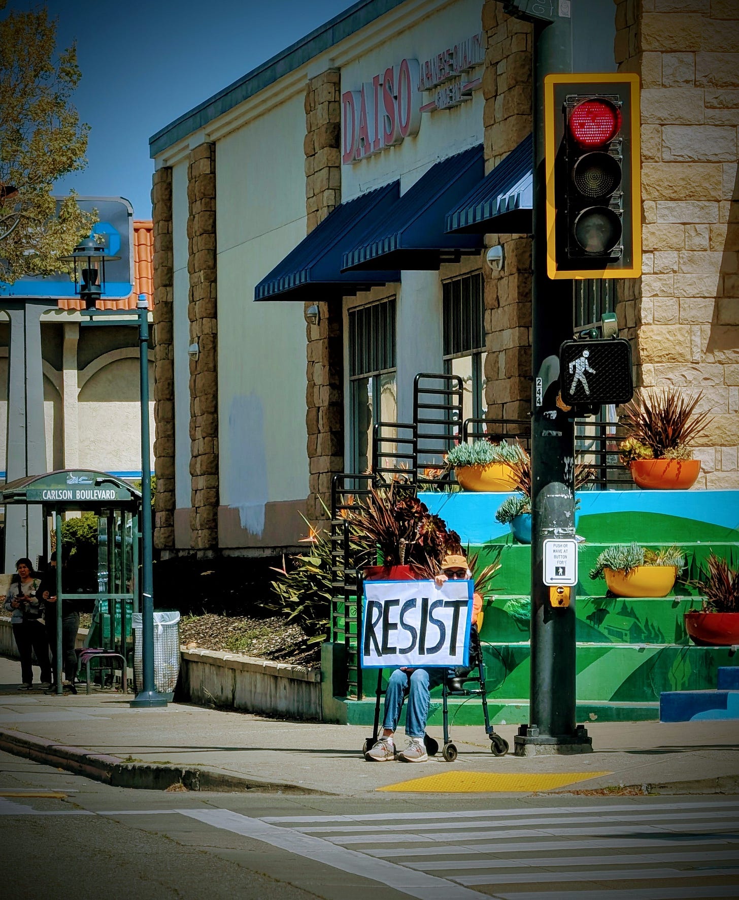 A person in a sunhat and sunglasses sits at a busy street corner in a folding chair, holding a handmade sign that reads “RESIST.” They sit alone beside a crosswalk, under a red light, near a bus stop and colorful planter boxes. The setting is everyday—but the message is unmistakable.