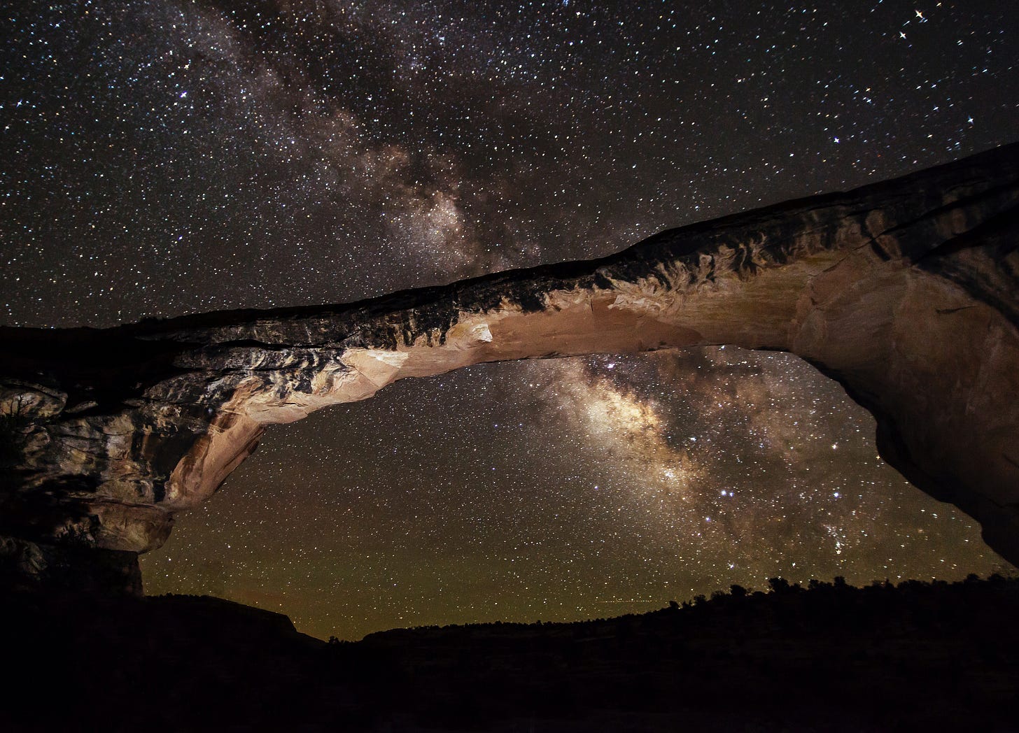 A bright band of stars crossed a dark sky full of stars with a geologic bridge formation in the foreground. A bright band of stars crossed a dark sky full of stars with a geologic bridge formation in the foreground.