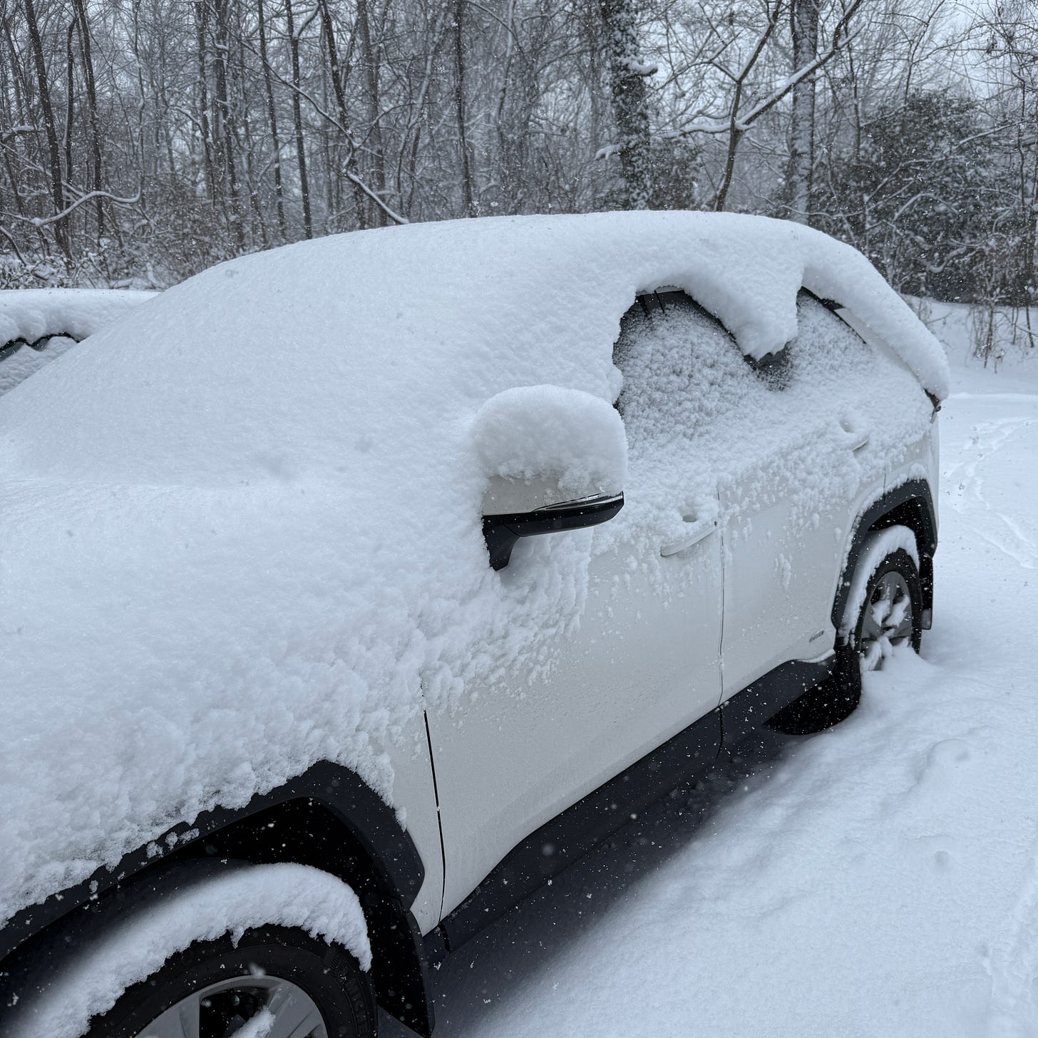 A white SUV sits in a driveway during a heavy North Carolina snowstorm, nearly buried under a thick blanket of fresh snow. Several inches have accumulated on the roof, hood, windows, and mirrors, while snow continues to fall among the leafless trees in the background. The vehicle appears almost sculpted from snow, illustrating why schools across the region shut down for nearly two weeks. A white SUV sits in a driveway during a heavy North Carolina snowstorm, nearly buried under a thick blanket of fresh snow. Several inches have accumulated on the roof, hood, windows, and mirrors, while snow continues to fall among the leafless trees in the background. The vehicle appears almost sculpted from snow, illustrating why schools across the region shut down for nearly two weeks.