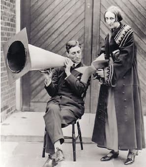 Edith Sitwell, with a friend holding a ‘sengerphone’, which looks like a large, possibly cardboard, traffic cone. Edith looks at the camera, and wears an elaborate robe. She is holding a manuscript for the young man, sitting and holding the device, to recite from