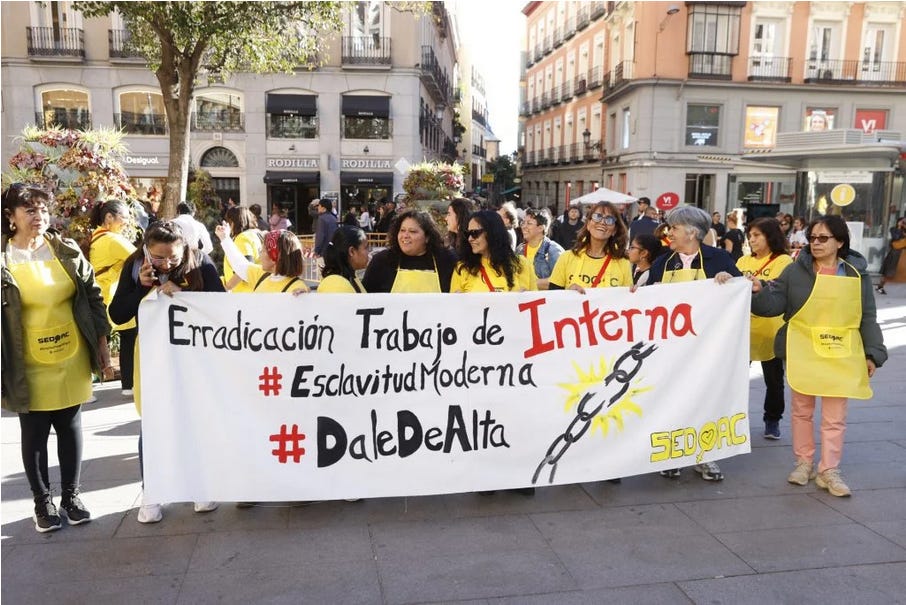 Un gruppo di donne del SEDOAC in una piazza di Madrid. Indossano un grembiule giallo con il nome dell’associazione e reggono uno striscione che dice: "Erradicación Trabajo de Interna. #EsclavitudModerna. #DaleDeAlta.” A fianco al testo, il disegno di una catena che si spezza. Un gruppo di donne del SEDOAC in una piazza di Madrid. Indossano un grembiule giallo con il nome dell’associazione e reggono uno striscione che dice: "Erradicación Trabajo de Interna. #EsclavitudModerna. #DaleDeAlta.” A fianco al testo, il disegno di una catena che si spezza.