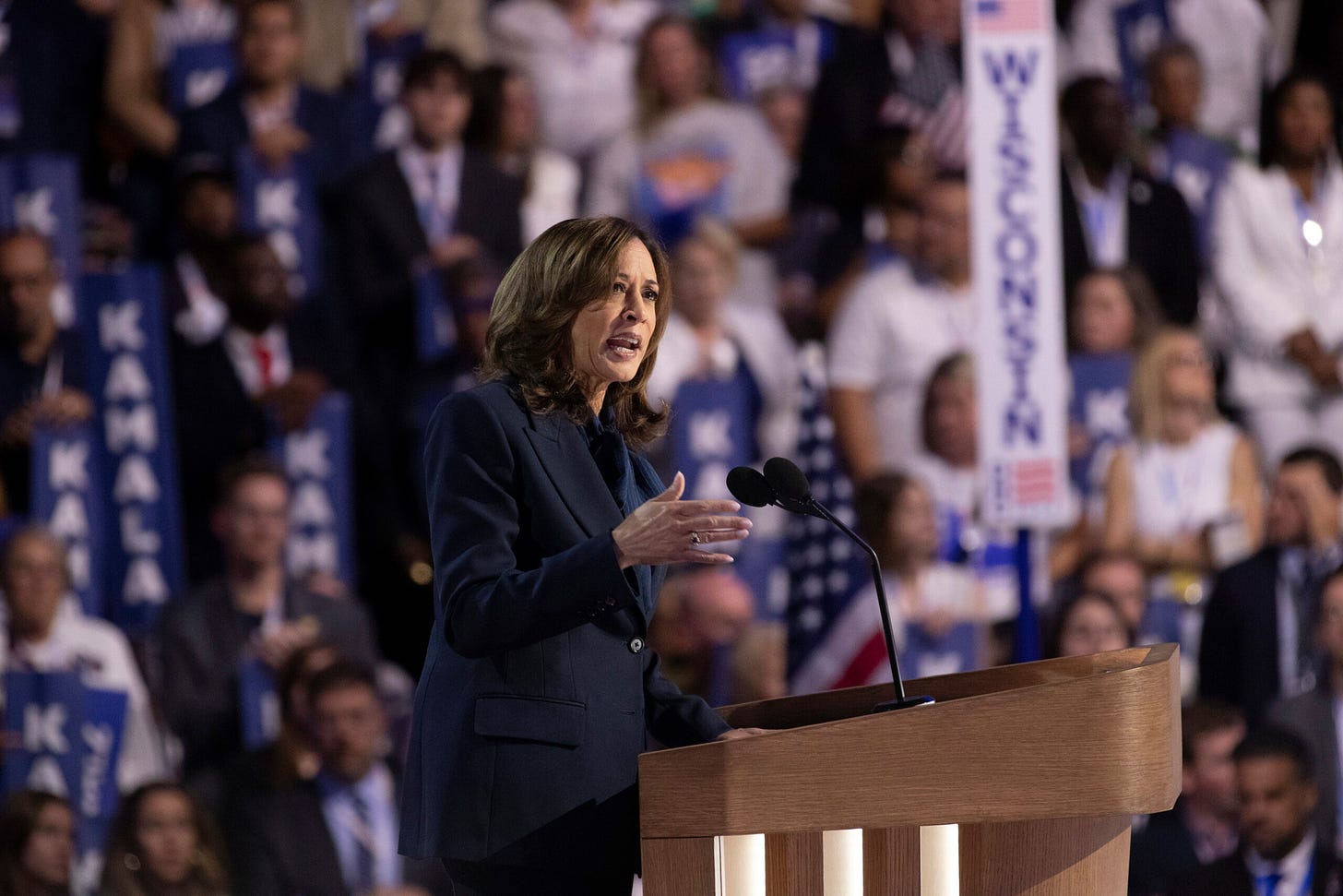 Kamala Harris is at a brown podium in the center of the frame, the DNC audience is behind her and blurred
