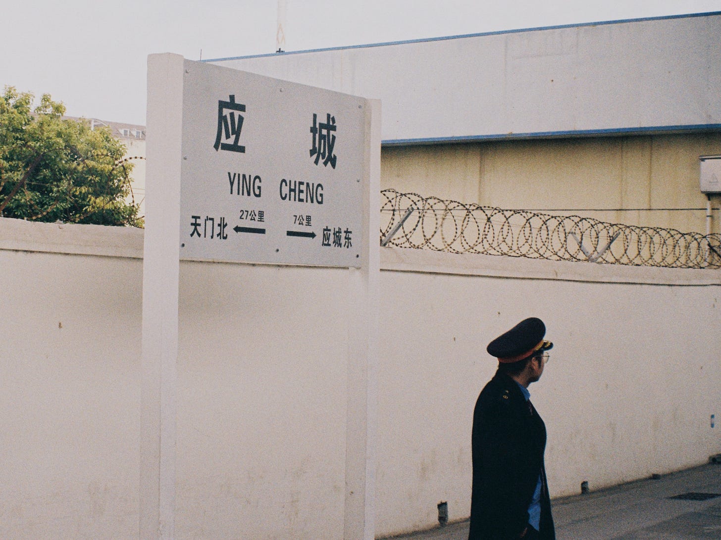 一张具有胶片质感的铁路纪实摄影。画面左侧是高大的白底黑字站牌，上面写着 Yingcheng (应城)，并标注了相邻站 Tianmen Bei (天门北) 和 Yingcheng Dong (应城东) 的距离。画面右侧，一名戴着大檐帽、身着深色制服的铁路工作人员正侧身站立，眺望着远方。背景是一堵带有铁丝网的白色围墙，整体氛围安静且带有怀旧色彩。A documentary-style railway photograph with a vintage film feel. On the left, a large white station sign displays Yingcheng (应城) in bold black characters, showing distances to neighboring stations Tianmen Bei (天门北) and Yingcheng Dong (应城东). On the right, a railway staff member in a dark uniform and peaked cap stands in profile, looking into the distance. The background features a white wall topped with barbed wire, creating a quiet and nostalgic atmosphere.