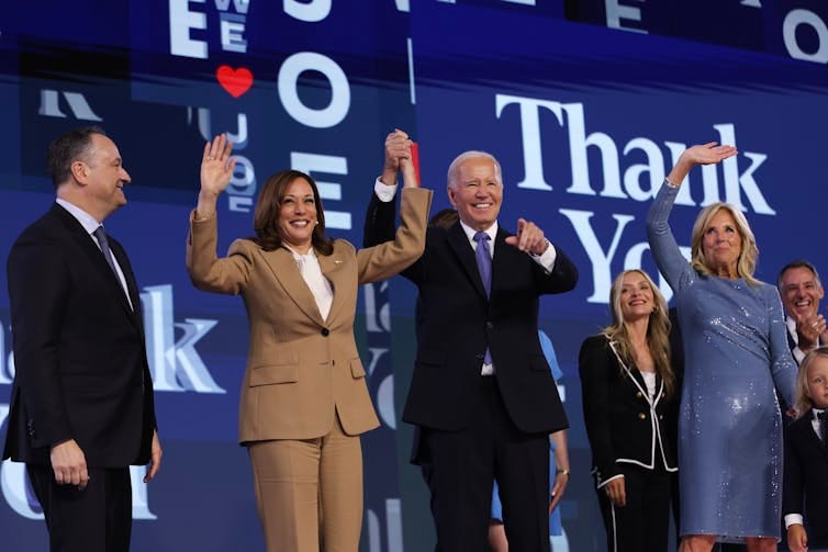 Four people standing on a stage, smiling and waving. Four people standing on a stage, smiling and waving.