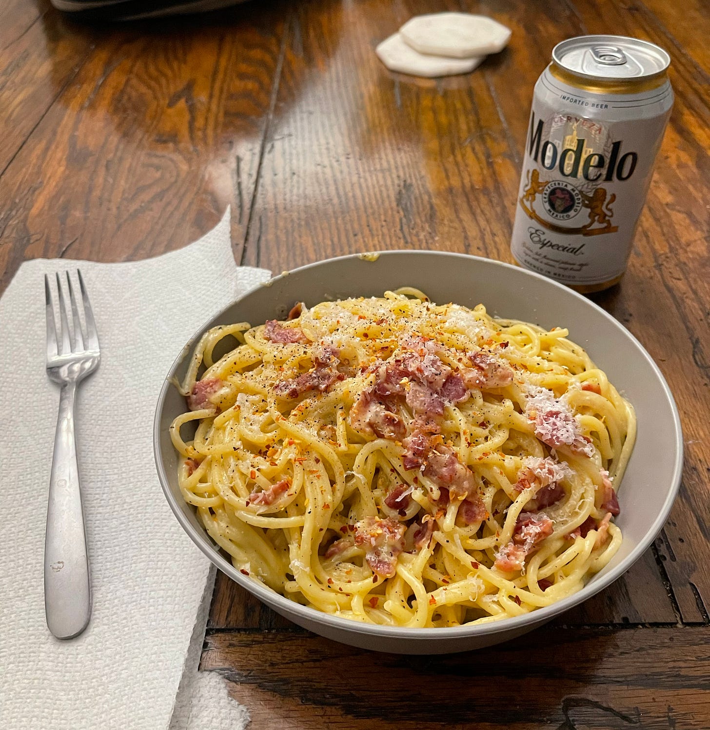 Bowl of homemade spaghetti carbonara that I made myself, plated in a wide plastic bowl on a wooden dining room table. A fork is sitting to the left side of the bowl on a piece of paper towel and a Modelo sits on the upper right side waiting to be enjoyed with the pasta.