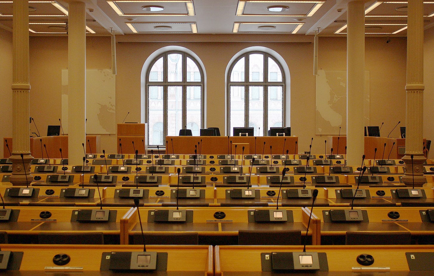 A photo of a city council room with rows and rows of desks with microphones at every seat facing the viewer while another desk at the far end faces in towards the others, in front of two large windows on the far wall.