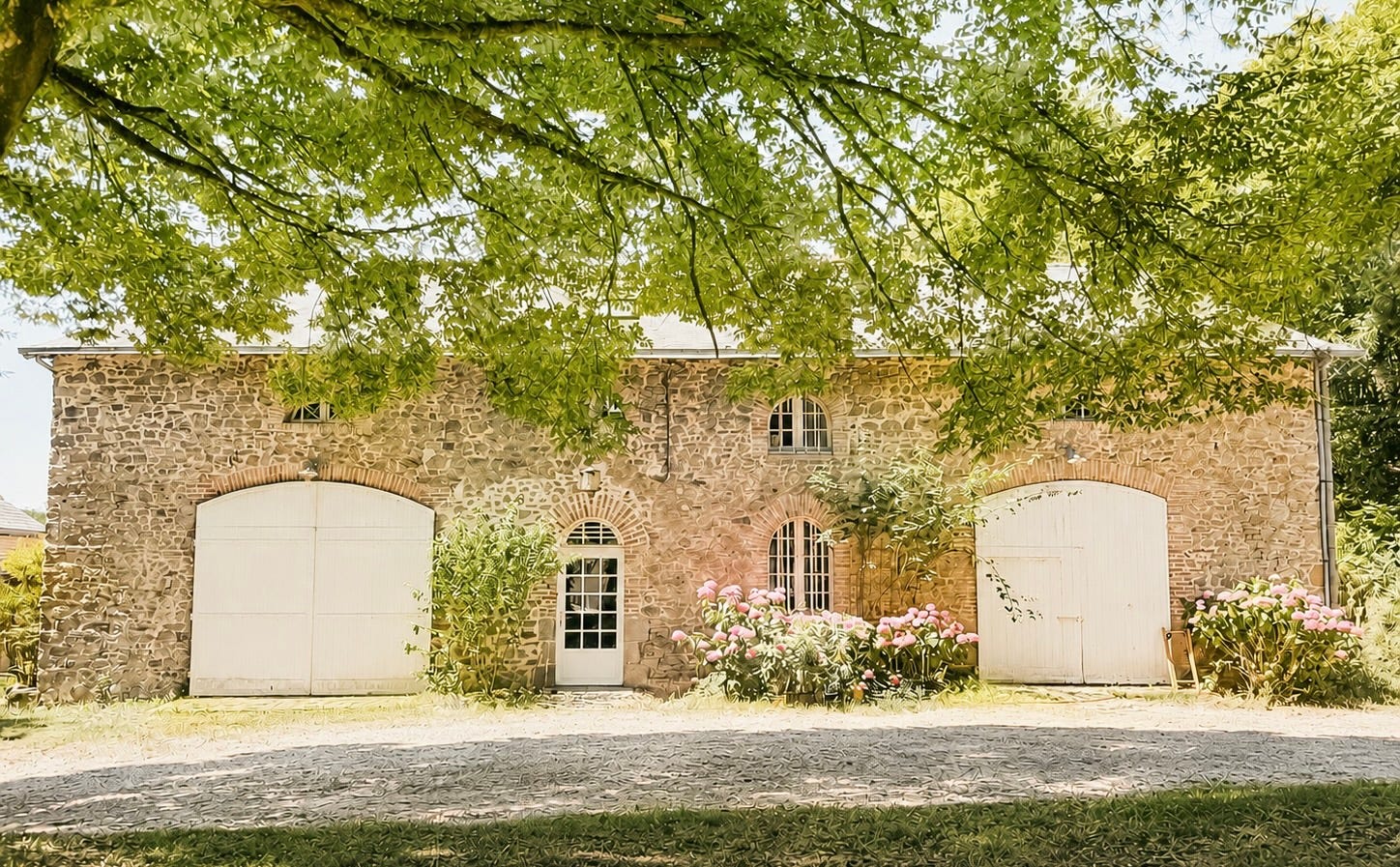 The front of a long stone coach house with two large white arched carriage doors, arched windows framed in brick, and pink hydrangeas blooming along the wall, seen beneath the leafy branches of a large tree on a sunny day. The front of a long stone coach house with two large white arched carriage doors, arched windows framed in brick, and pink hydrangeas blooming along the wall, seen beneath the leafy branches of a large tree on a sunny day.