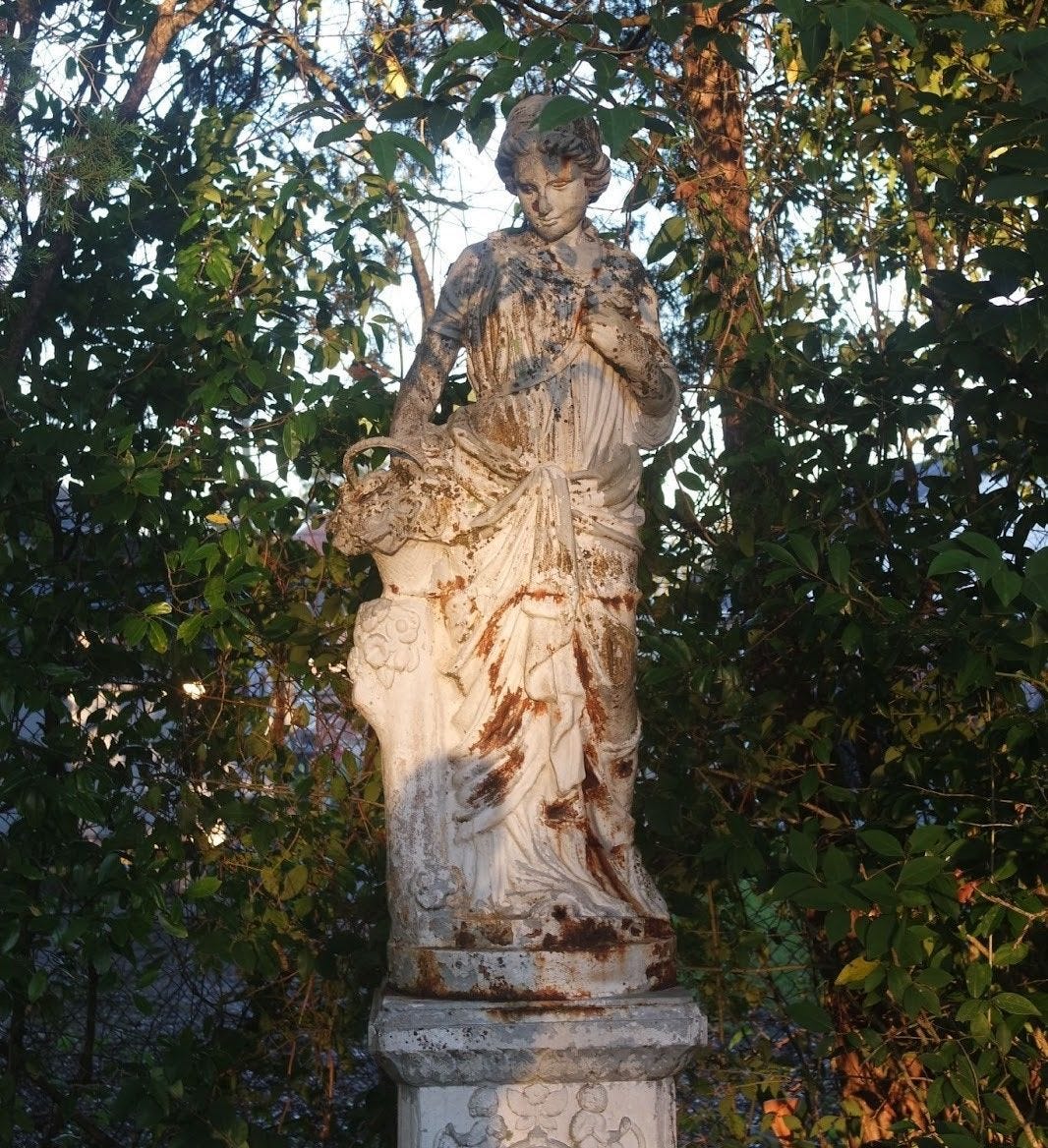 A weathered classical stone statue of a draped female figure stands on a pedestal amid overgrown vines and foliage.