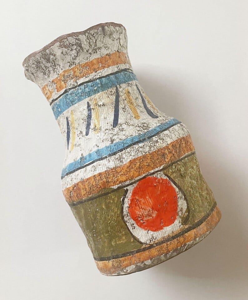May include: A handcrafted, textured vase with a unique, aged appearance. The vase features a white base with horizontal bands of orange and blue, accented by vertical blue and yellow lines. A green band with a red circle detail adds a pop of color.