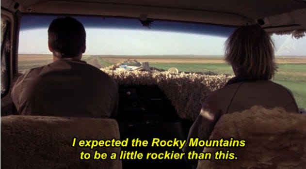 two men driving in a van with backs turned, window looks out at a flat boring scenery of beige fields