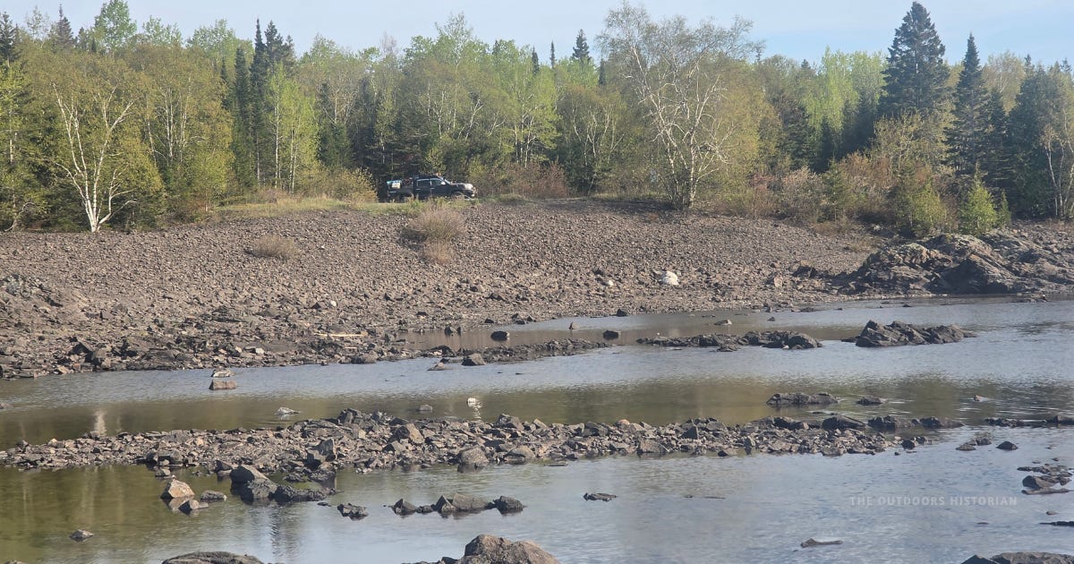View from shallow water looking back toward shore, where a Toyota Tacoma is parked in the distance under a bright blue sky. Rocks and calm Lake Superior water fill the foreground.