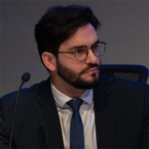 Homem adulto, de pele clara, cabelo escuro e barba, usando óculos, terno escuro, camisa branca e gravata azul. Ele está em um ambiente que lembra uma audiência ou reunião formal, com microfone à frente.