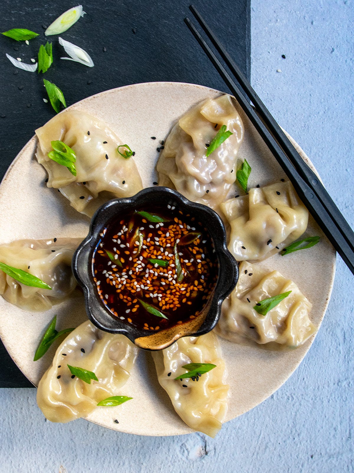 Korean mandu on a tan plate surrounding a brown bowl filled with dipping sauce.