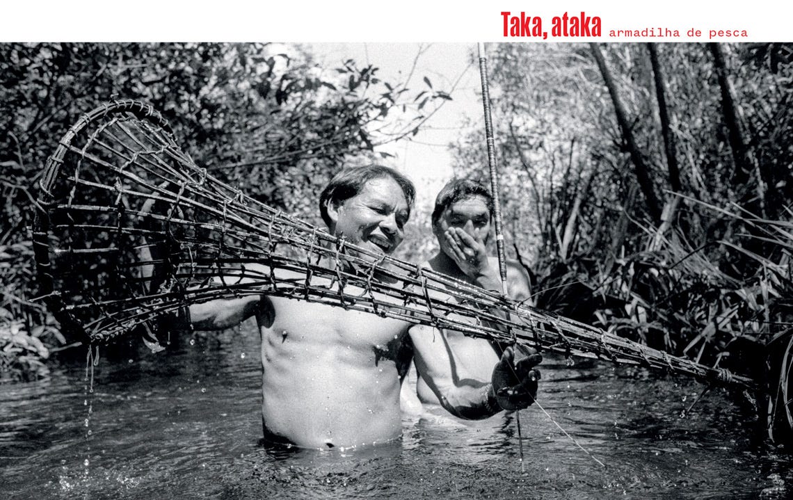 #descrevemospravocê  Taka, ataka – armadilha de pesca.  A imagem em preto e branco mostra dois homens adultos, de pele clara, em meio a um igarapé raso, com água na altura da cintura, cercados por vegetação alta e densa nas margens. O homem à frente sorri amplamente enquanto segura uma armadilha de pesca tradicional em forma de funil alongado, feita de madeira e cipós, utilizada para capturar peixes. Ele está de corpo nu, com gotas d’água escorrendo pela armadilha que acabou de retirar da água. Atrás dele, o segundo homem observa, com uma das mãos tapando parte do rosto e um leve sorriso, demonstrando cumplicidade e descontração. O cenário é de floresta, com galhos e folhas refletindo a luz do sol entre sombras, transmitindo a atmosfera de prática tradicional de pesca em ambiente de rio ou igarapé na floresta, ressaltando a cultura e os saberes locais sobre técnicas de pesca artesanal.