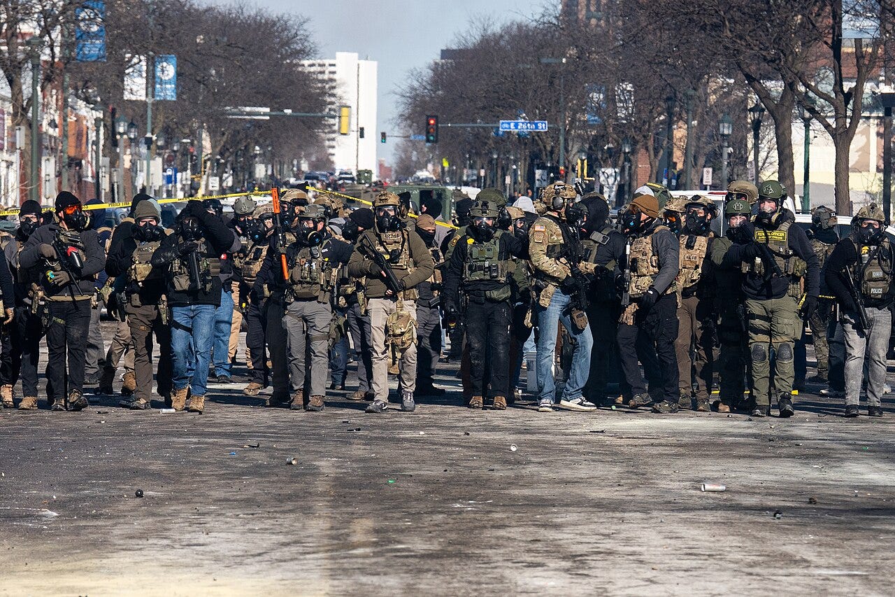 A dense line of men with gas masks, body armor, and rifle block a wide city street in winter. A dense line of men with gas masks, body armor, and rifle block a wide city street in winter.