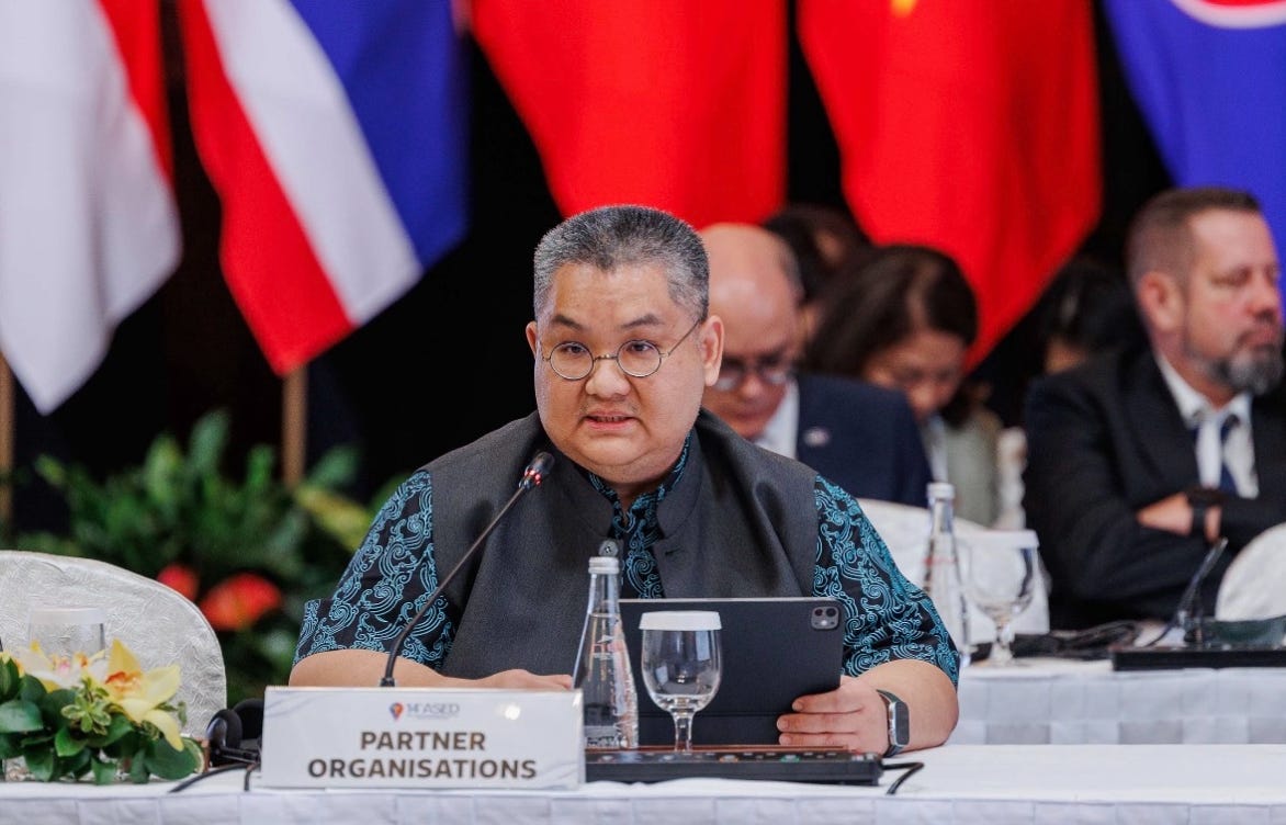 Dr. Piti Srisangnam, wearing glasses, a dark vest, and a blue patterned shirt, speaks into a microphone at a conference table. In front of him are a drinking glass, a tablet, and a nameplate reading "PARTNER ORGANISATIONS", set against a backdrop of national flags. Dr. Piti Srisangnam, wearing glasses, a dark vest, and a blue patterned shirt, speaks into a microphone at a conference table. In front of him are a drinking glass, a tablet, and a nameplate reading "PARTNER ORGANISATIONS", set against a backdrop of national flags.