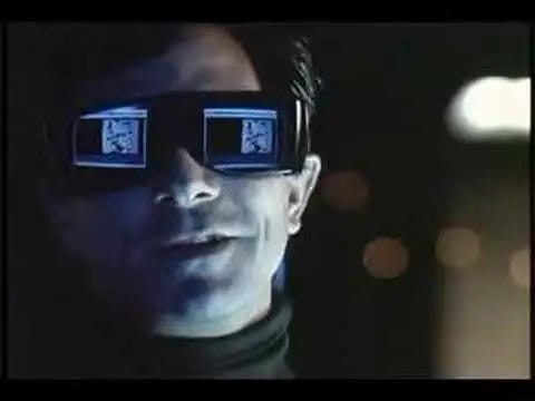 A film still of David Straithairn playing the blind hacker character "whistler" in Sneakers. He's wearing big sunglasses with the command line reflected hugely in each lens.