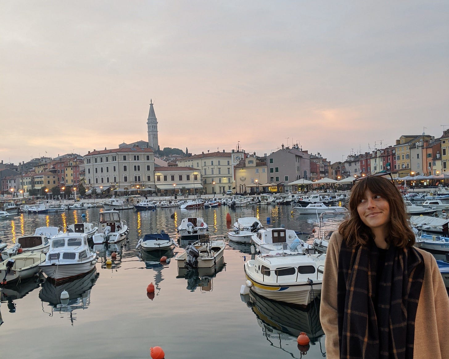A woman stands by a marina at dusk, smiling slightly as small boats float on calm water behind her, with pastel-coloured buildings and a church tower lining the waterfront.