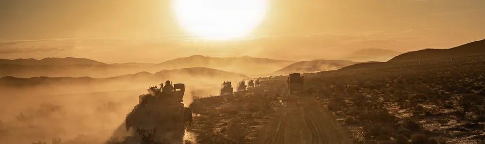 A military convoy of armored and wheeled vehicles travels along a dirt road through an arid desert landscape at either sunrise or sunset. The vehicles are kicking up significant dust clouds as they move in a column formation toward the horizon. The scene is dramatically backlit by a large, bright sun low on the horizon, creating silhouettes and a golden haze across the terrain. A military convoy of armored and wheeled vehicles travels along a dirt road through an arid desert landscape at either sunrise or sunset. The vehicles are kicking up significant dust clouds as they move in a column formation toward the horizon. The scene is dramatically backlit by a large, bright sun low on the horizon, creating silhouettes and a golden haze across the terrain.