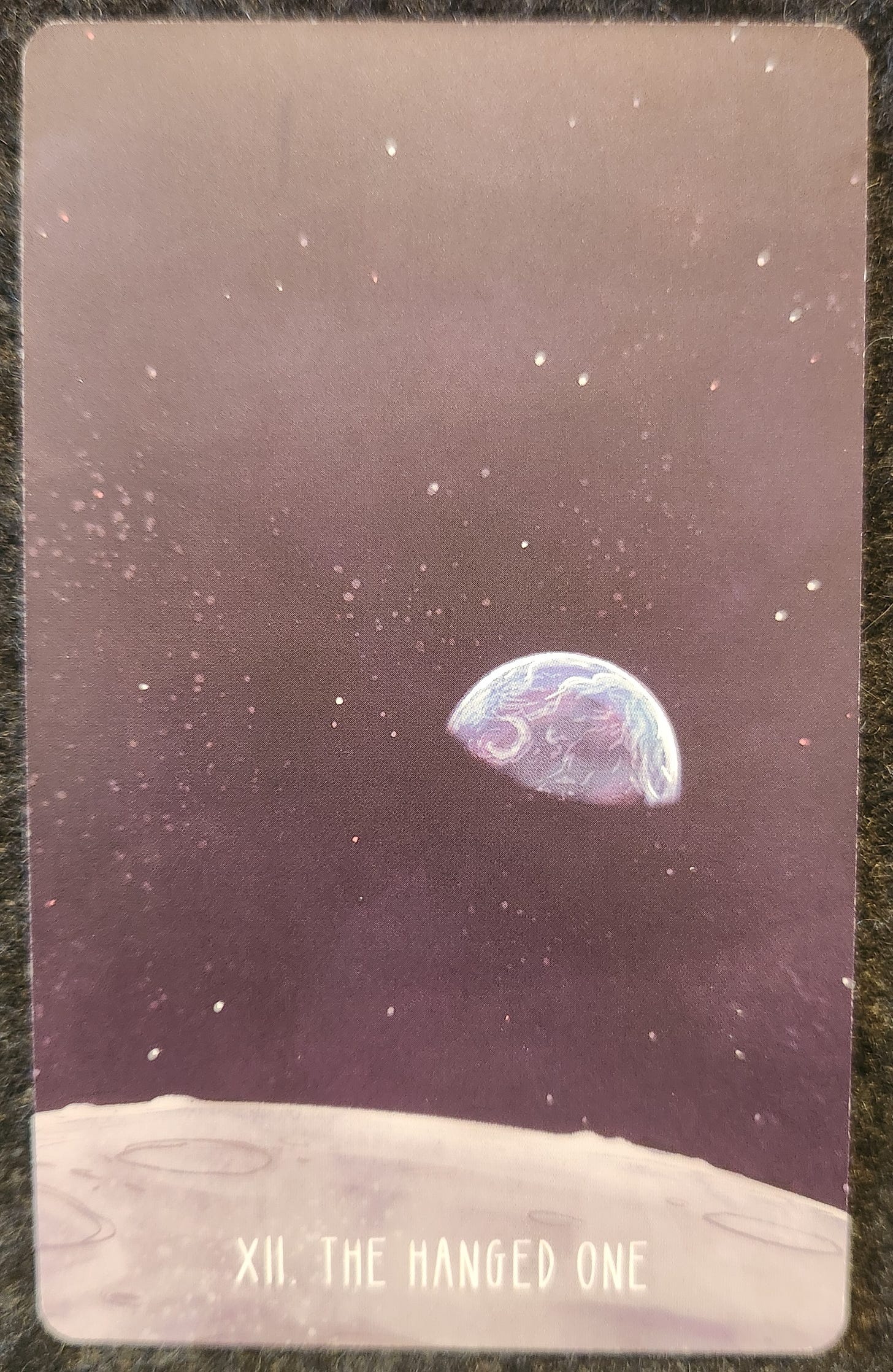 A tarot card labelled XII The Hanged One. A view of the Earth from outer space, as though from the perspective of standing on the moon. The Earth is half covered in darkness. The sky is black with many visible stars. 