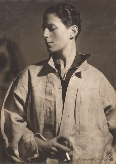 1) Classic black-and-white portrait of Gluck in a loose white shirt/smock, looking down and to the side. Their expression is thoughtful, with shadows adding depth. They have short dark hair, and they are holding a cigarette in their right hand. There is a dark shirt sticking out from under the larger shirt, with both collars sticking up. 2) A white mannequin displays a vintage, cream-colored linen shirt/smock with a black collar and buttoned cuffs, evoking a classic, timeless style. It's positioned in the same style Gluck was wearing it in the previous photo, including the collars sticking up. The mannequin head is also looking to the side, like Gluck was. 3) A pair of vintage brown leather shoes with buckles and low heels on a neutral background, conveying a sense of timeless style and craftsmanship. 4) Five vintage dresses/gowns hang on a black clothing rack with a small jacket. Patterns include floral, paisley, and abstract designs, evoking a nostalgic and elegant feel. 5) A white mannequin displays an elegant black evening gown with a lace overlay. The dress features long sleeves and a floor-length skirt, conveying sophistication.