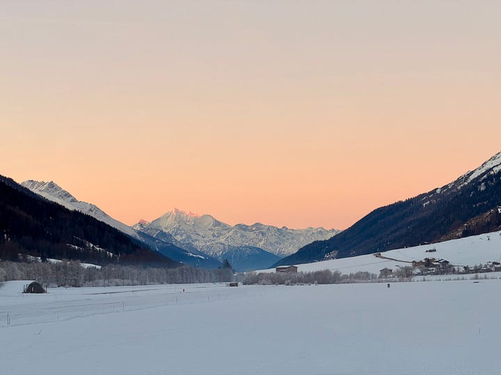 Left image, A snow-covered alpine valley plateau at sunrise. The peak of the centered Weisshorn mountain is sun kissed, and the sky around it tinted pink. The snow of the valley, still in shadows has a bluish tint; right image, the sun sets behind a mountain slope as view from a snow-covered valley plateau. Two walkers make their way along a groomed path in the foreground. In the background, the sharp, snow-covered Weisshorn mountain. The setting sun flashes a golden aura and tints the clouds above the mountains and snow on the valley floor orange.