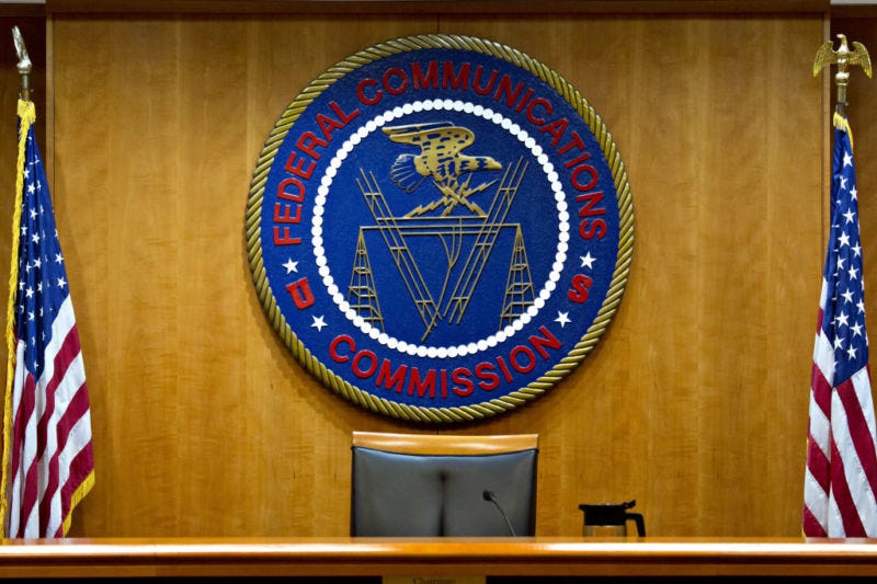 The Federal Communications Commission meeting room, with an empty chair in front of the FCC seal and two United States flags.