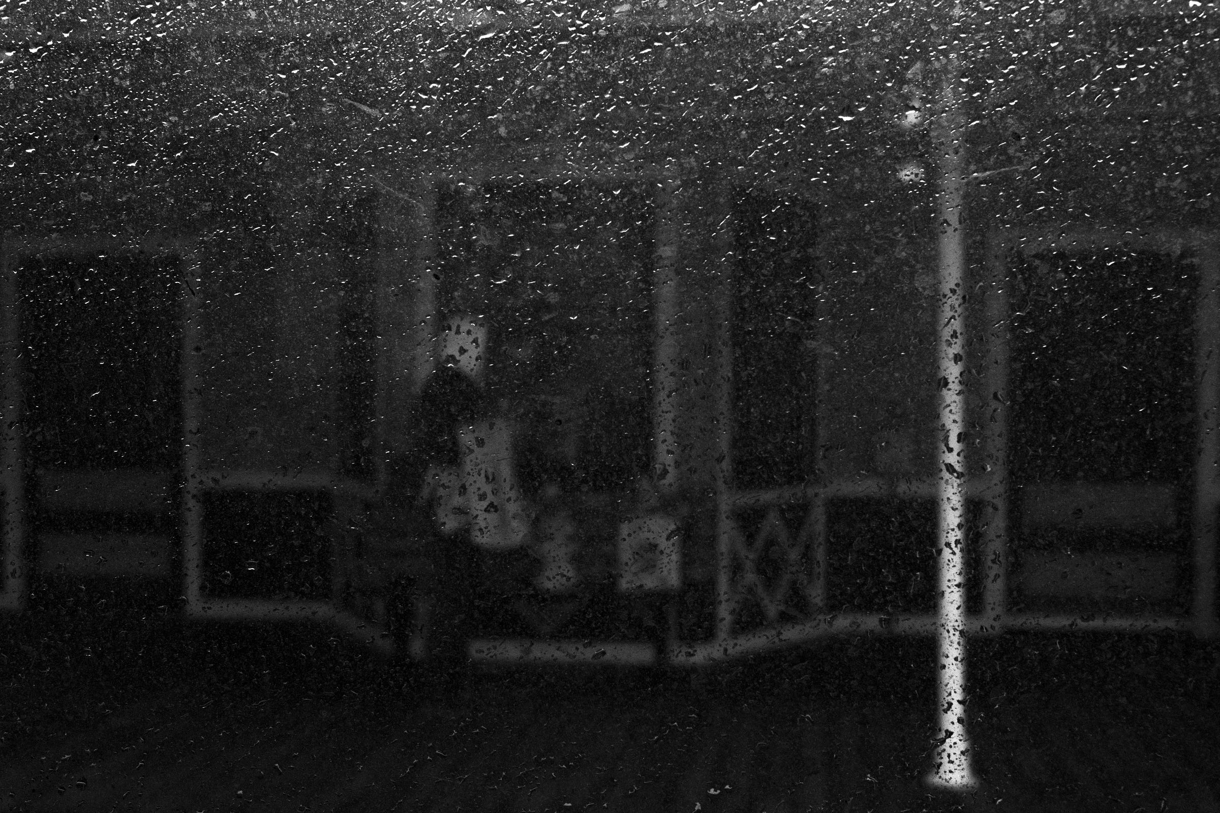 Rain-speckled window obscuring the view of a person sitting on a porch, with a white pillar and railing faintly visible in the background. Rain-speckled window obscuring the view of a person sitting on a porch, with a white pillar and railing faintly visible in the background.