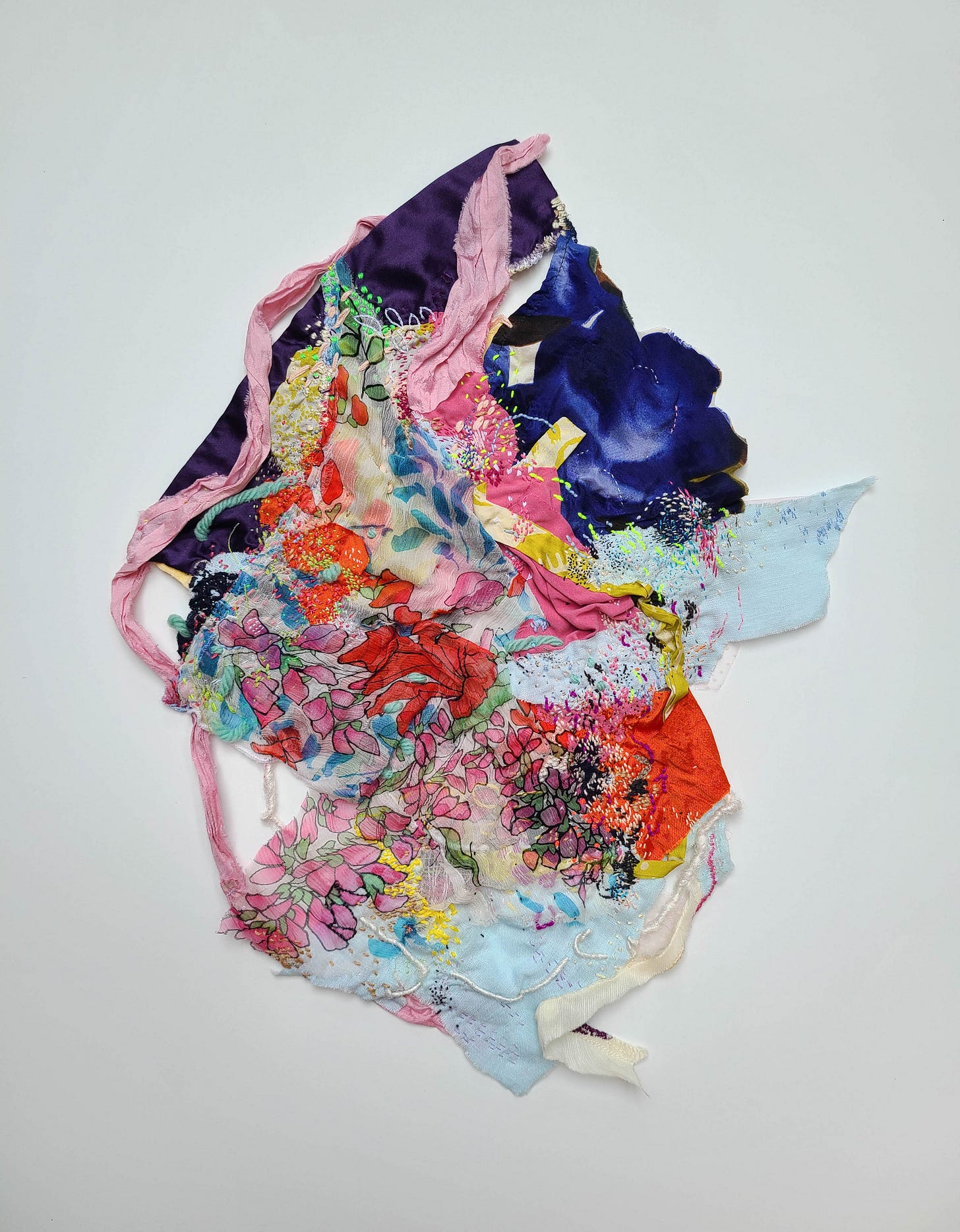 An abstract textile art piece composed of various colorful fabric scraps and embellishments. The piece features a mix of textures, patterns, and embroidery in shades of purple, pink, blue, orange, and yellow, creating a vibrant and chaotic composition.