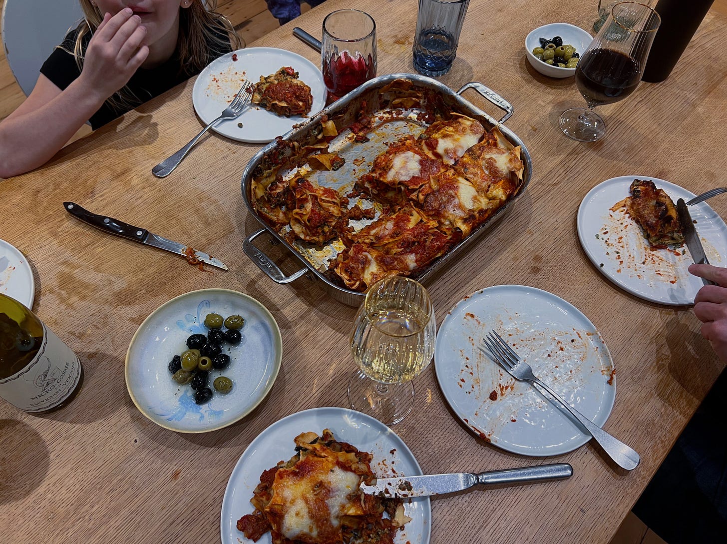 A table set with a pan of lasagna, half of which has been served, surrounded by plates with lasagna eaten in varying degrees, a plate of olives, and glasses of wine, and a kids elbow on the table with their hand to their mouth.