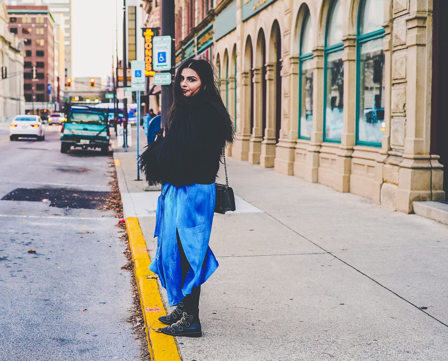 a person wearing a blue skirt and fluffy jacket, with long black hair and Doc Martin boots, is standing with their arms crossed and a perturbed look on their face. They are standing on a footpath in a busy looking city.