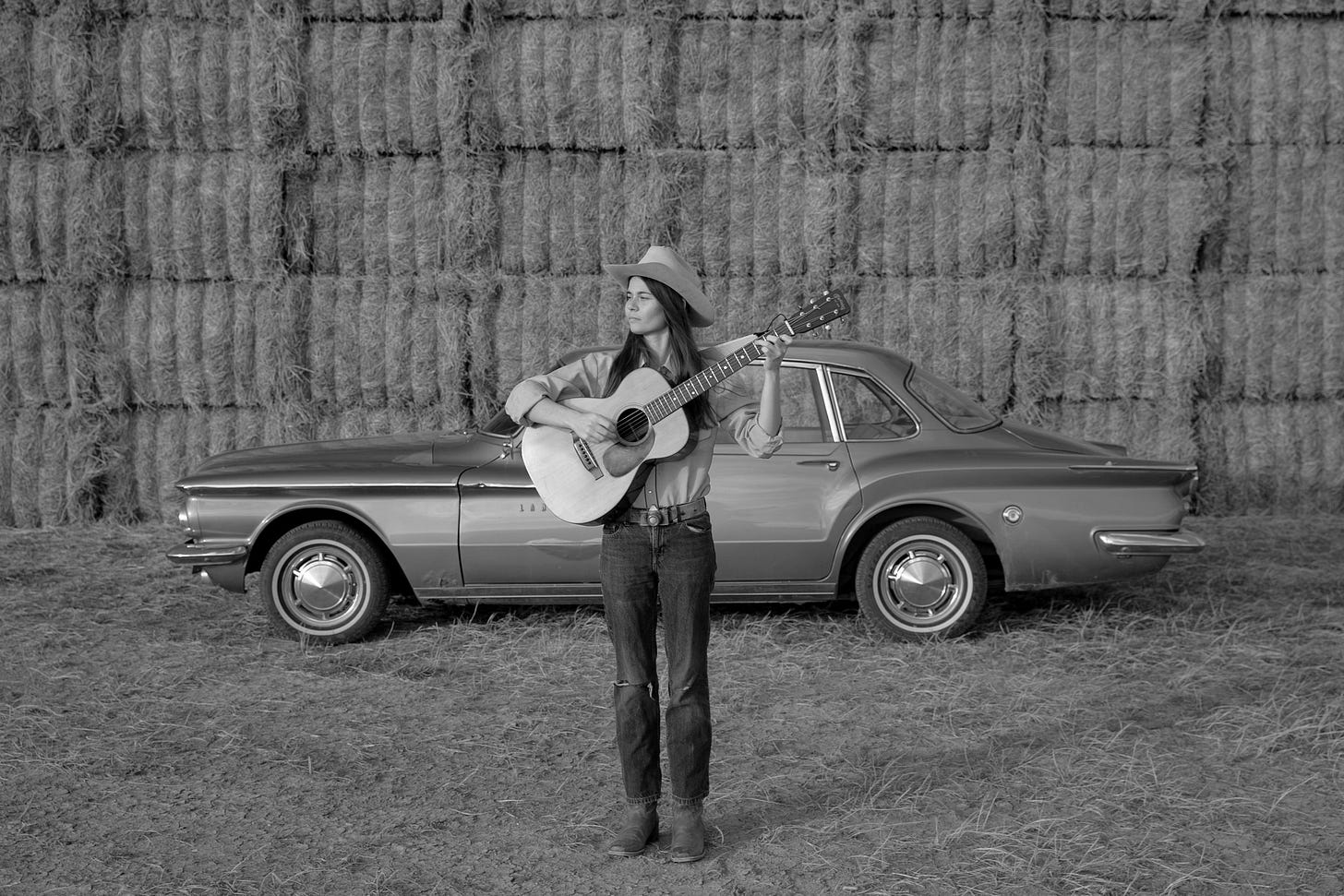 black white photo of woman with guitar in front of car and bales of hay