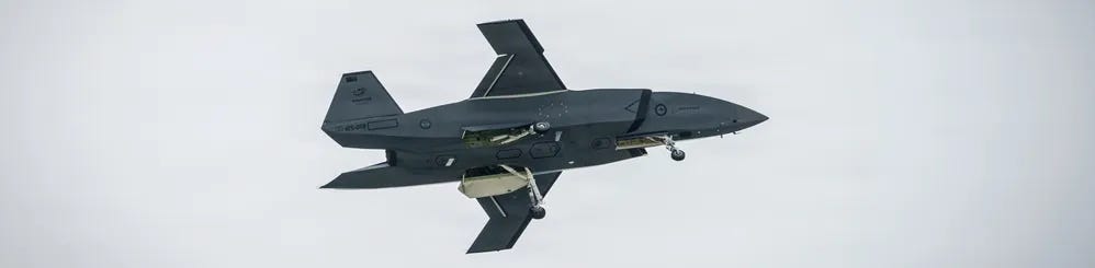 A dark-colored unmanned aerial vehicle (UAV) or drone aircraft is captured in flight against an overcast gray sky. The aircraft appears to be in a banking maneuver with landing gear deployed, suggesting it may be on approach or during a test flight. Markings visible on the fuselage suggest a military affiliation, consistent with the Boeing MQ-28 Ghost Bat or similar advanced combat drone. A dark-colored unmanned aerial vehicle (UAV) or drone aircraft is captured in flight against an overcast gray sky. The aircraft appears to be in a banking maneuver with landing gear deployed, suggesting it may be on approach or during a test flight. Markings visible on the fuselage suggest a military affiliation, consistent with the Boeing MQ-28 Ghost Bat or similar advanced combat drone.