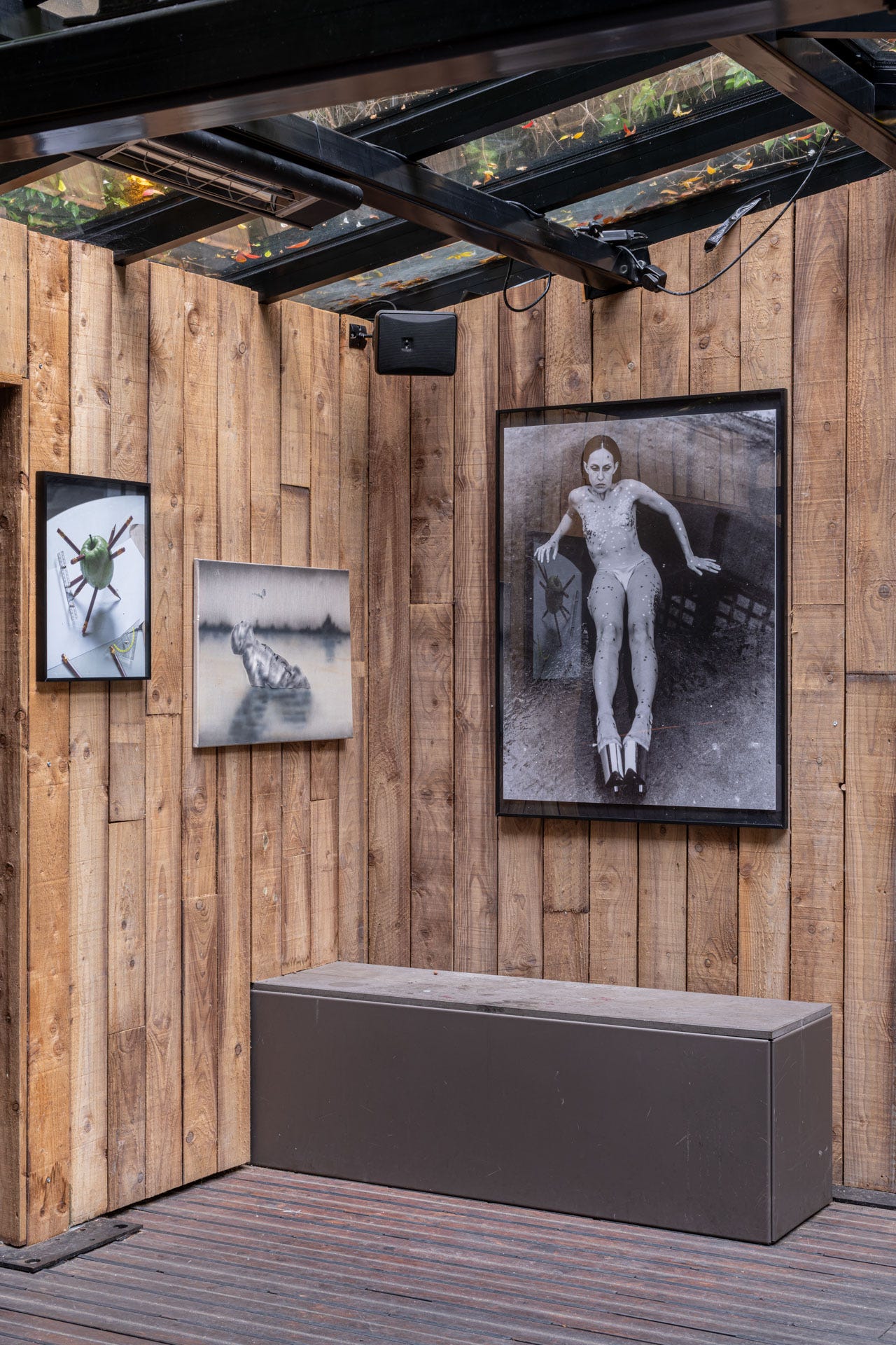 Wood-paneled interior with framed artworks hung salon-style beneath a dark ceiling grid, creating a rustic, intimate presentation.