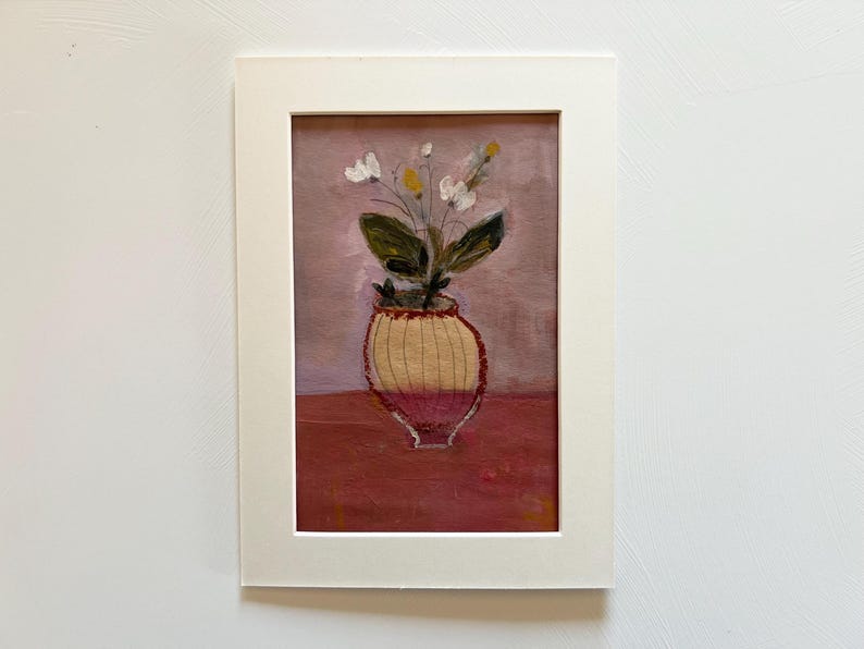 May include: An original painting of a vase with flowers, set in a white matted frame. The vase is yellow and red, with green leaves and white and yellow flowers. The background is a muted pink and red.