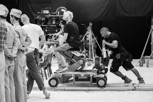 A black-and-white image shows a man sitting on a complicated rig with a movie camera. Another man is leaning his whole body in to push the rig.