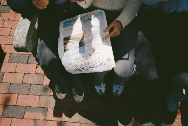 An overhead photo of an issue of The Ditch Weekly seemingly shared by two young readers. It rests between their laps.