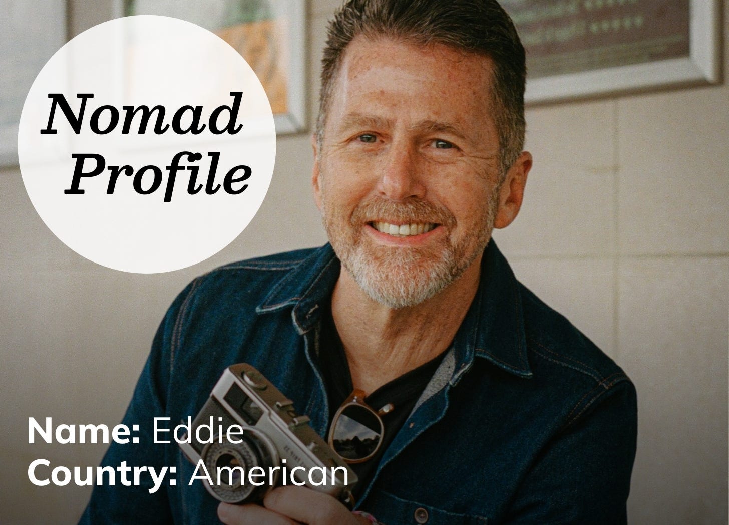 White man with a grey beard is wearing a blue-jean shirt and smiling. He is holding a vintage camera and has sunglasses hanging from his collar. 