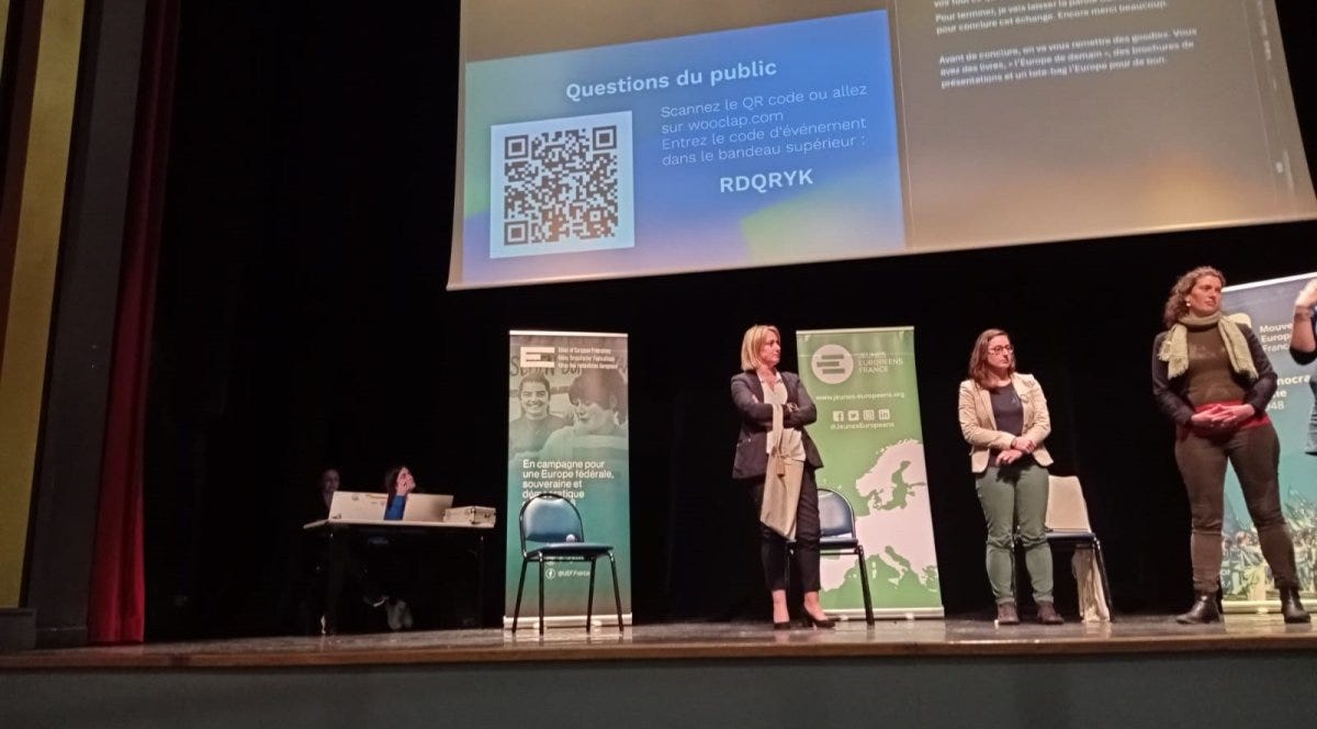 Sur une scène, trois personnes débattent  devant un grand écran. Au fond, Céline est derrière son ordinateur.