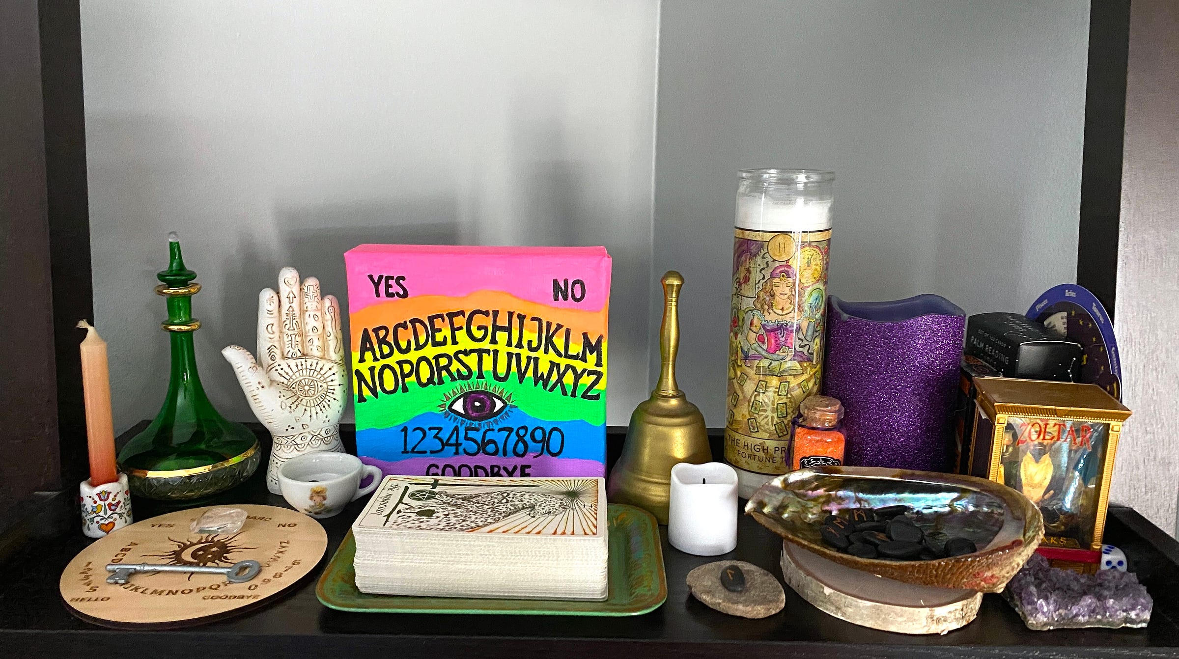 A shelf on a bookshelf in the author’s home, featuring a painting designed to look like a rainbow Ouija board, the Wild Unknown Tarot deck (card facing up: The Magician), an abalone shell filled with runestones, a bell, candles, a glass bottle, and a miniature Zoltar fortune telling machine.
