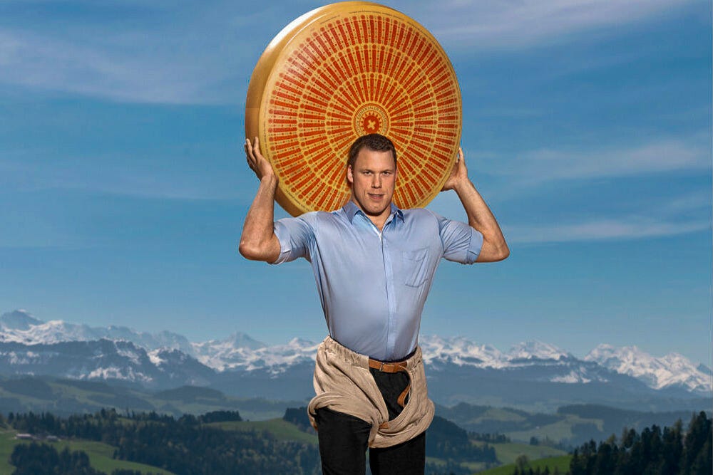 Schwingerkönig Matthias Sempach steht vor einem Panorama aus grünen Hügeln und schneebedeckten Alpen und hält einen grossen, runden Emmentaler-Käselaib kraftvoll über beiden Schultern. Er trägt ein blaues Hemd und eine spezielle Überhose aus Zwilch. Schwingerkönig Matthias Sempach steht vor einem Panorama aus grünen Hügeln und schneebedeckten Alpen und hält einen grossen, runden Emmentaler-Käselaib kraftvoll über beiden Schultern. Er trägt ein blaues Hemd und eine spezielle Überhose aus Zwilch.